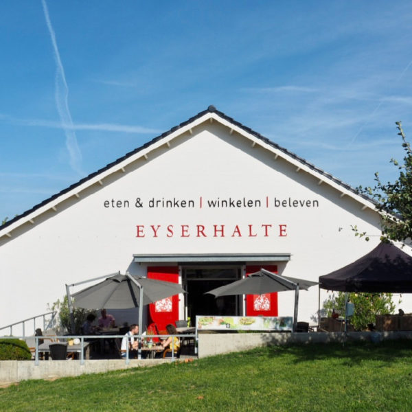 Vooraanzicht van lunchwinkel en delicatessenzaak de Eyserhalte. Een witte gevel met rode deuren en terras ervoor.